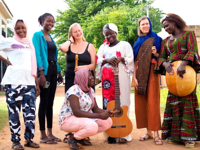 Five Malian women musicians and two Finnish women musicians are standing on the sand street in Bamako, Mali.
